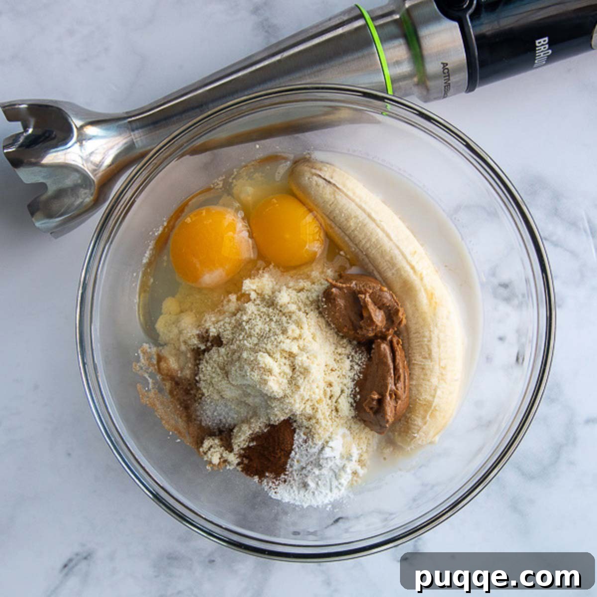 All the ingredients for almond flour banana pancakes are placed in a glass bowl, with an immersion blender resting beside it, ready for blending into a smooth batter.