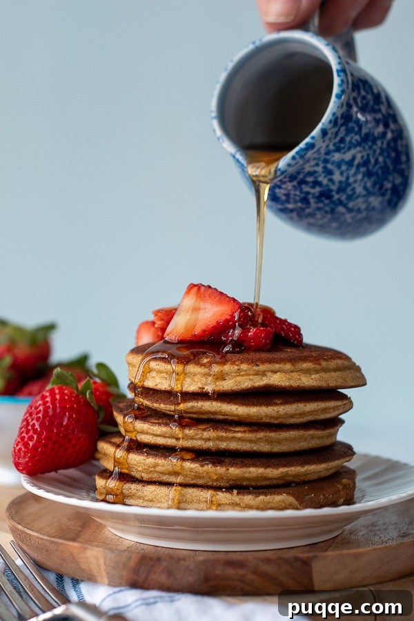 A generous stream of golden maple syrup being poured over a high stack of perfectly cooked almond flour pancakes, creating a luxurious and enticing visual for breakfast.