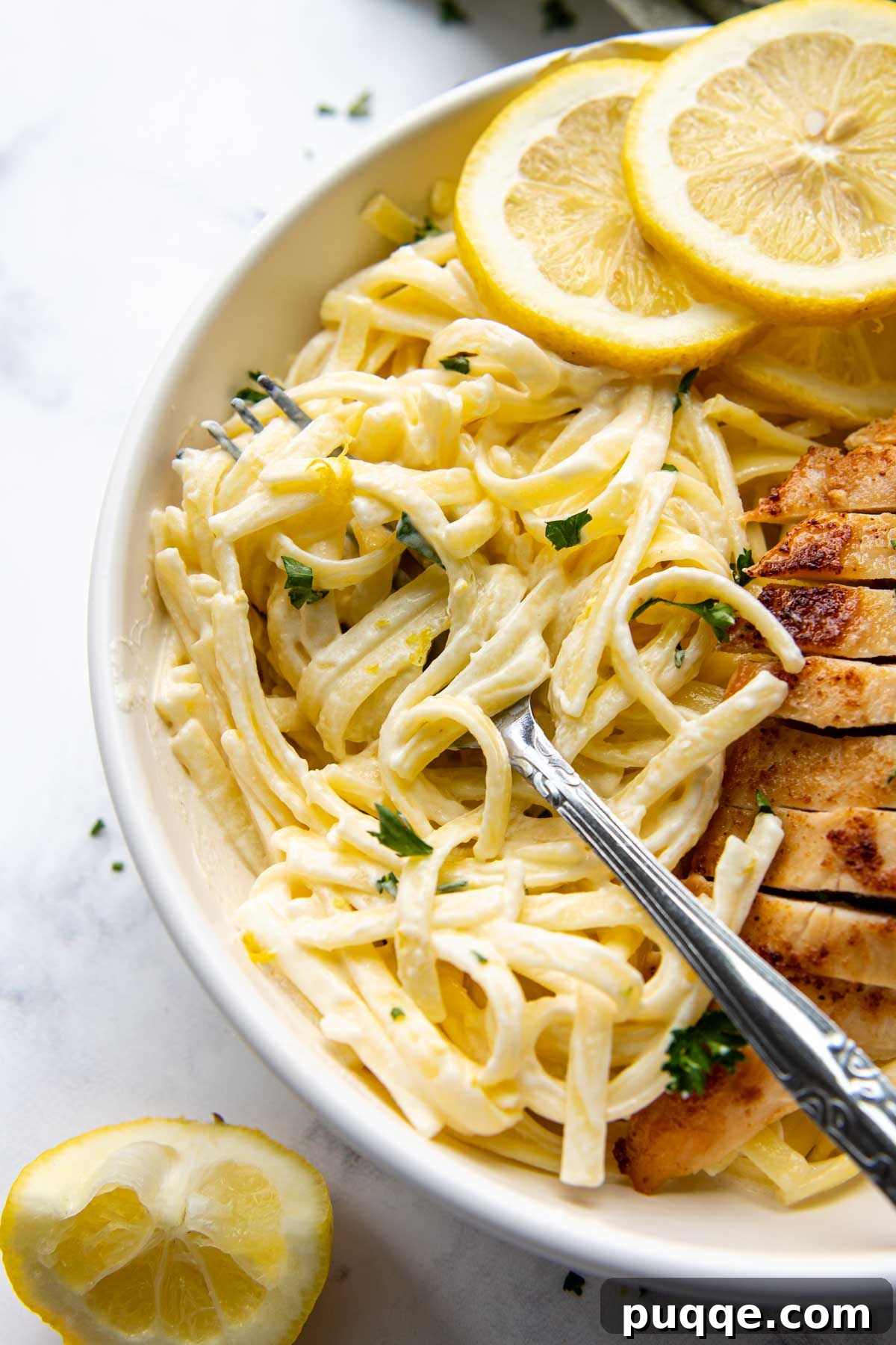 A fork piercing pasta in a bowl of creamy lemon chicken pasta, garnished with fresh lemon slices.