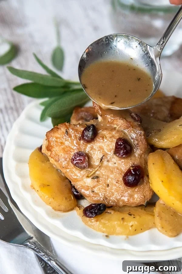 Pork medallions on a white plate, served with apples and cranberries, with a ladle of creamy Dijon gravy being poured over.