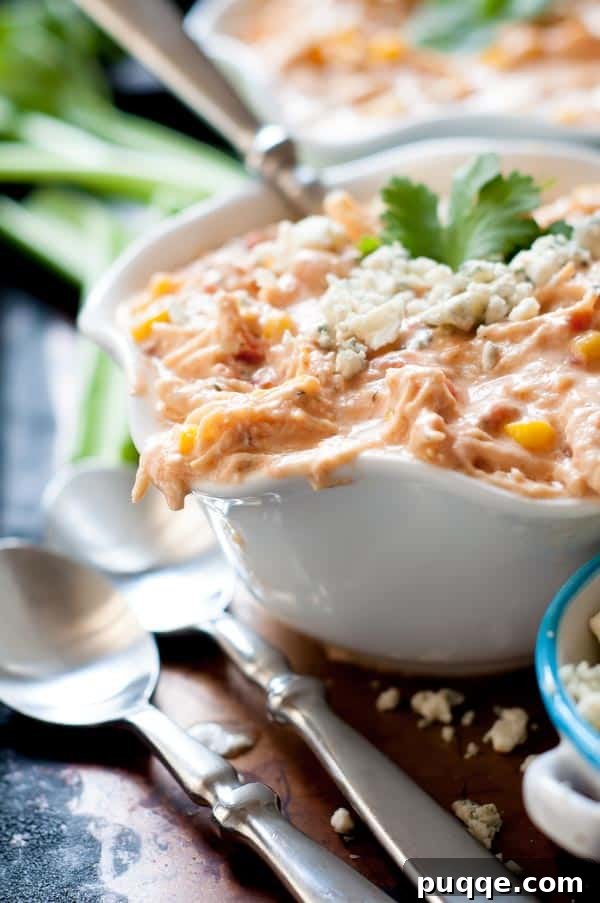 Award Winning Slow Cooker Buffalo Chicken Chili 2 A close-up shot of a bowl of creamy buffalo chicken chili, garnished with a sprinkle of fresh herbs. Two spoons rest beside the bowl, inviting a taste.
