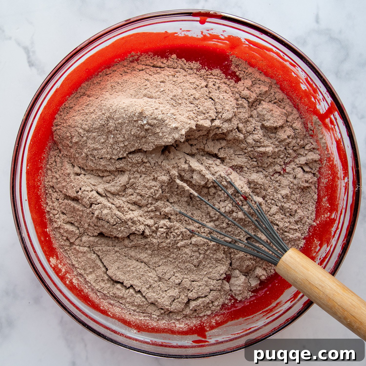 Decadent Gluten Free Red Velvet Pure Moist Perfection 4 Dry ingredients like flour and cocoa powder being gently whisked into the red liquid batter in a glass bowl, showcasing the mixing process.