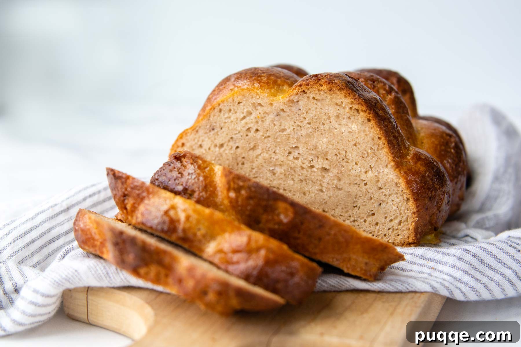 Baking Gluten Free Challah Made Easy 11 A half-sliced loaf of golden gluten-free challah bread resting on a wooden cutting board with white linen underneath, showcasing its tender interior.