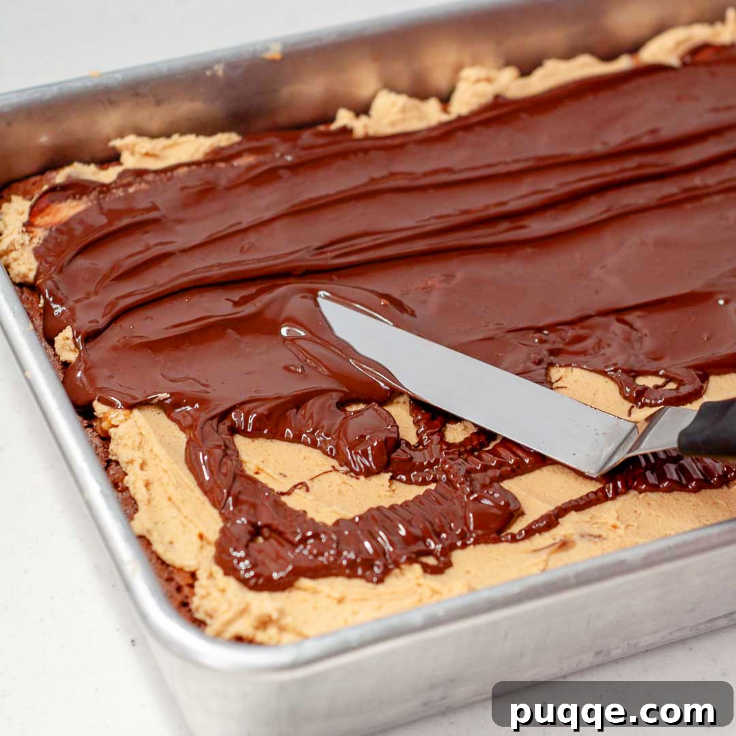 A pristine layer of melted chocolate being smoothly spread over the creamy peanut butter topping, forming the final decadent layer of the Buckeye Turtle Brownies.