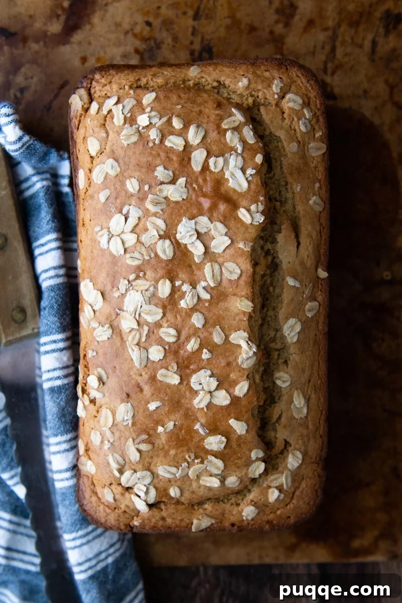 overhead shot of oat flour bread with oats sprinkled on top, a blue towel, and a knife next to it, ready to be sliced