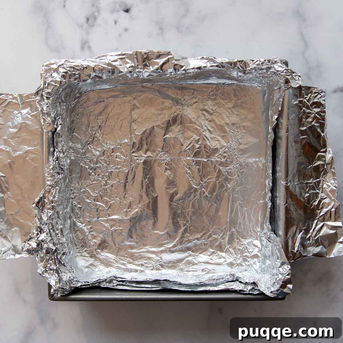 An empty 8x8 inch baking pan lined with two overlapping sheets of aluminum foil, creating a sling for easy removal.