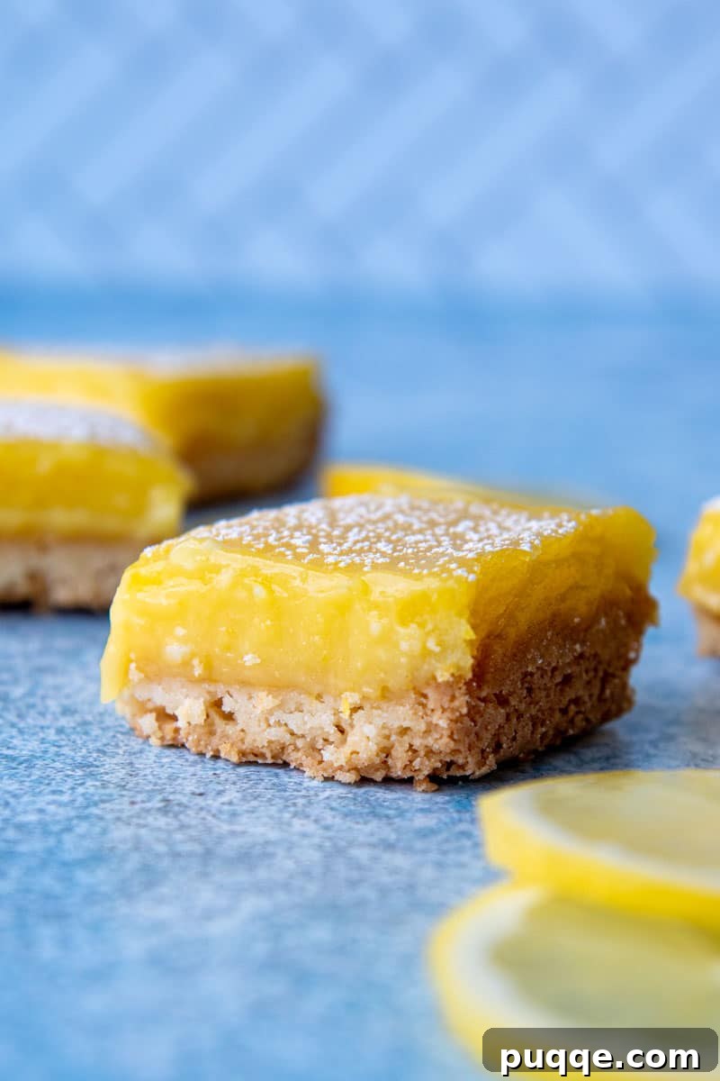 A single cut gluten-free lemon square sitting on a blue cutting board, with more lemon bars visible in the blurred background.