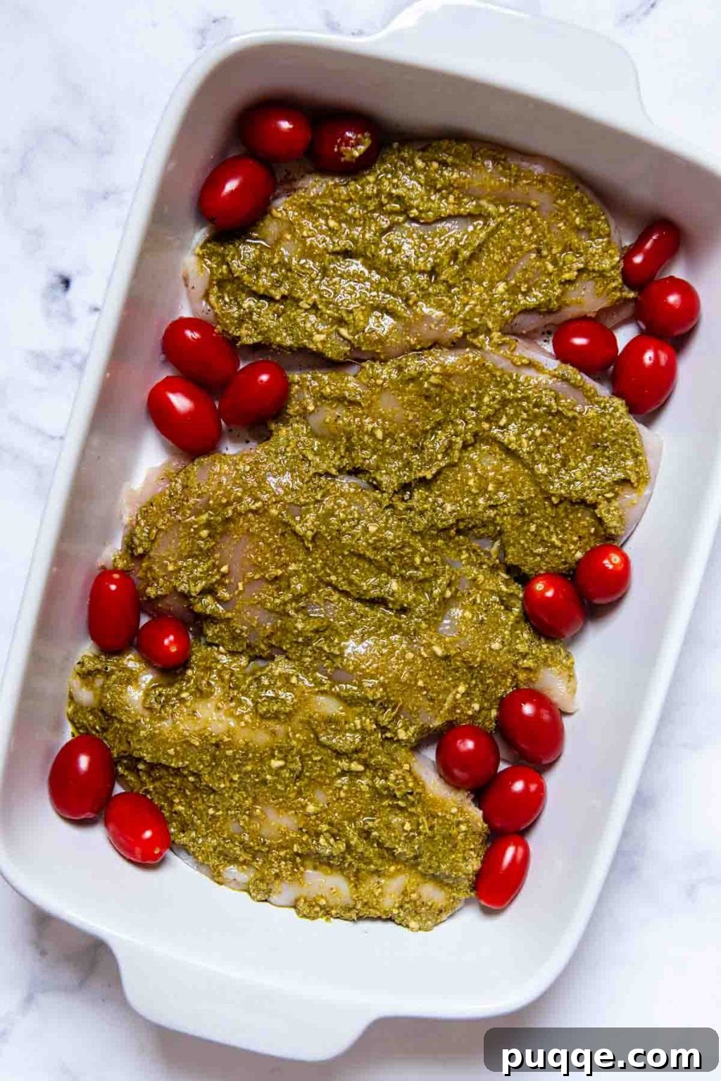 An uncooked chicken breast in a white baking dish, generously coated with vibrant green pesto, surrounded by bright red cherry tomatoes, ready for baking.