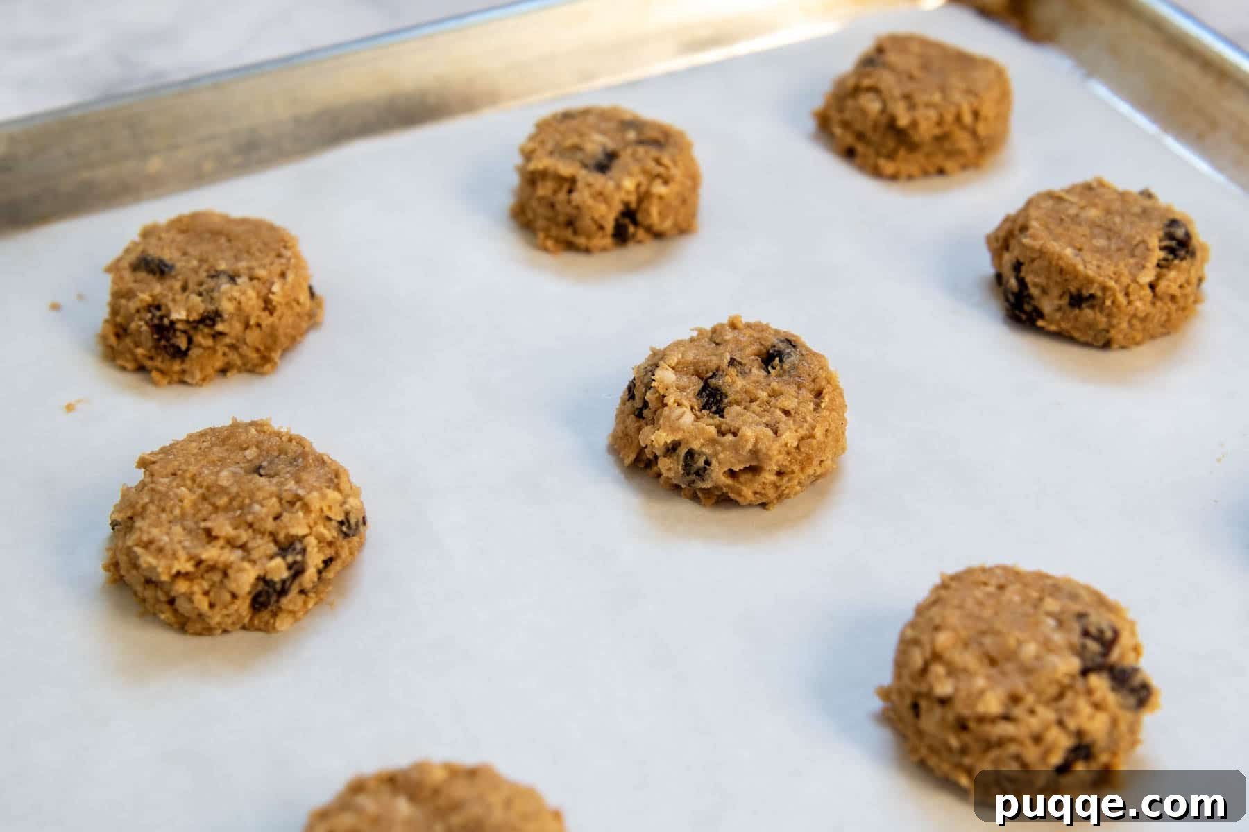 15 Minute Gluten Free Flourless Oatmeal Raisin Cookies 4 Unbaked gluten-free oatmeal raisin cookie dough balls neatly arranged on a parchment-lined cookie sheet, ready for baking.