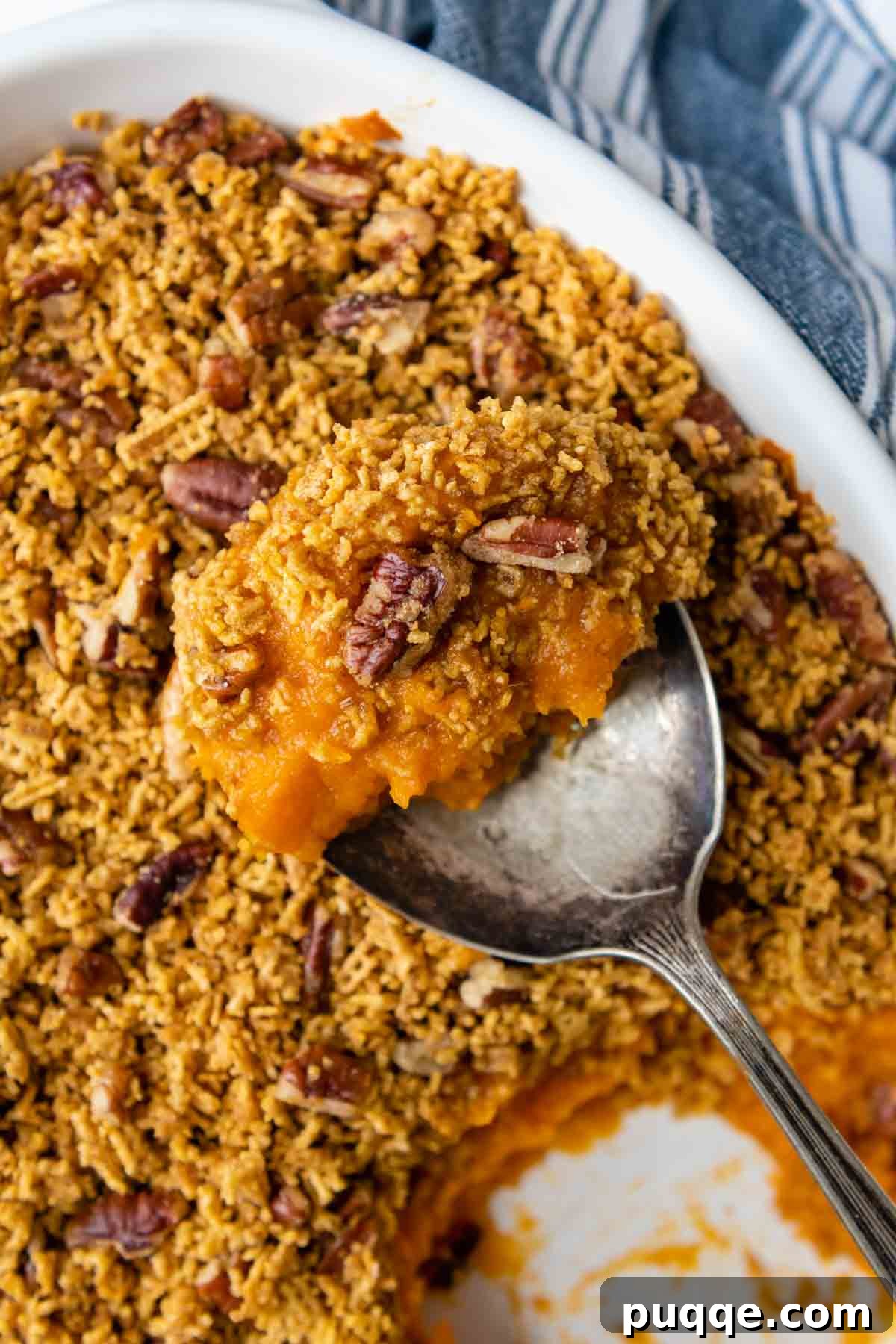 A serving spoon gracefully lifting a portion of Crunchy Sweet Potato Casserole from a baking dish, revealing its fluffy interior and irresistible pecan Chex topping.