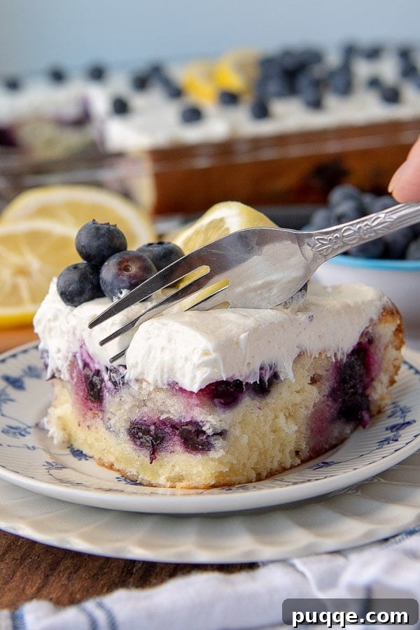 A fork poised to cut into a beautiful slice of lemon blueberry cake on a pristine white plate, garnished with fresh blueberries on top, inviting a delightful taste.