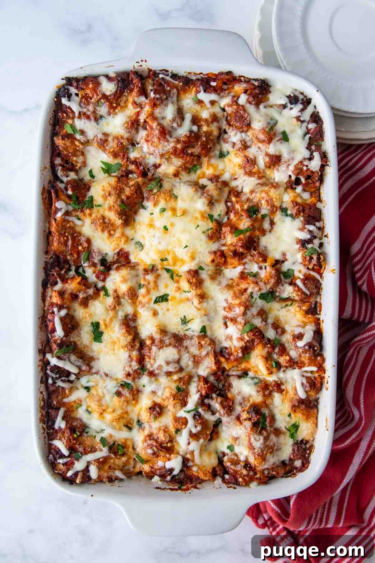An overhead shot of a golden-brown lasagna in a white baking dish, with a red kitchen towel beside it.