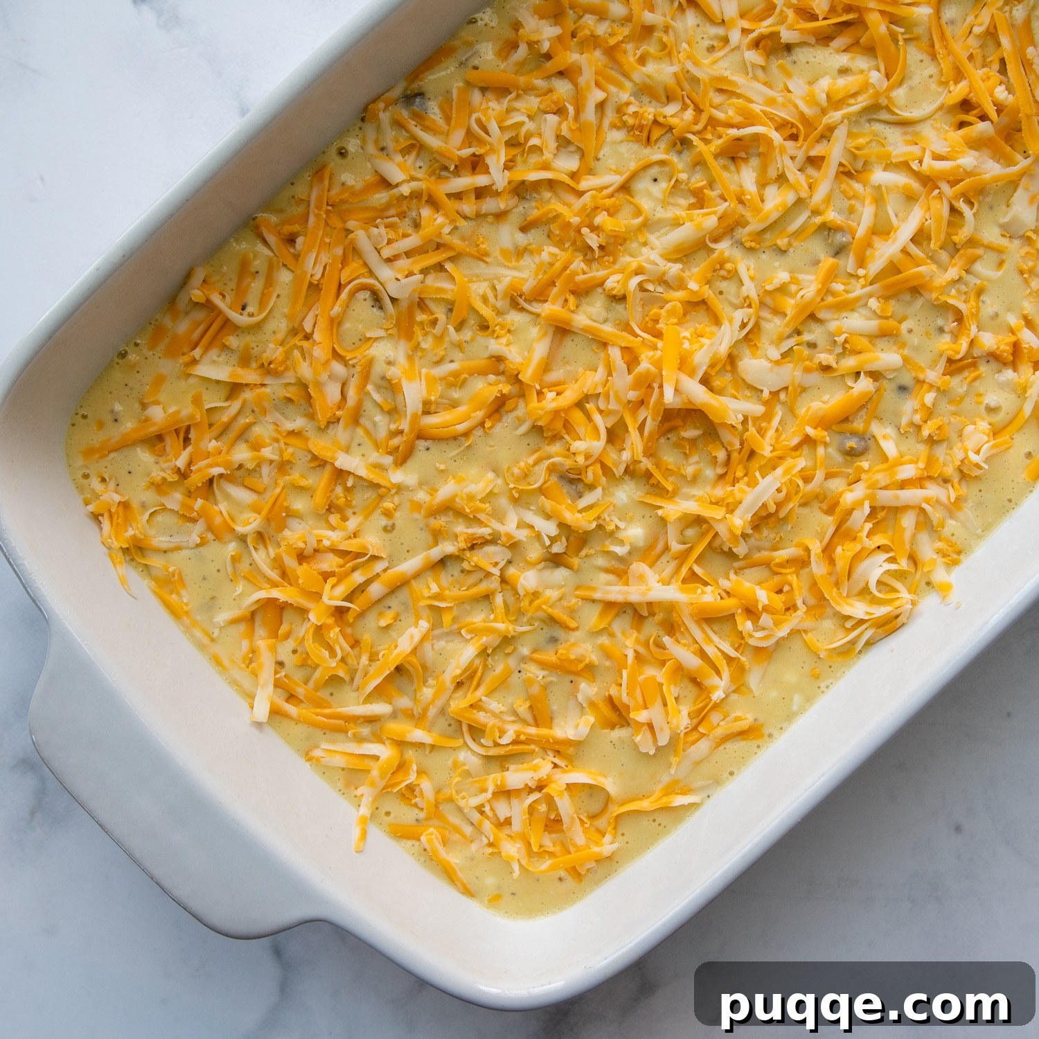 The egg green chile casserole mixture in a baking dish, generously sprinkled with extra cheese on top, ready for baking.