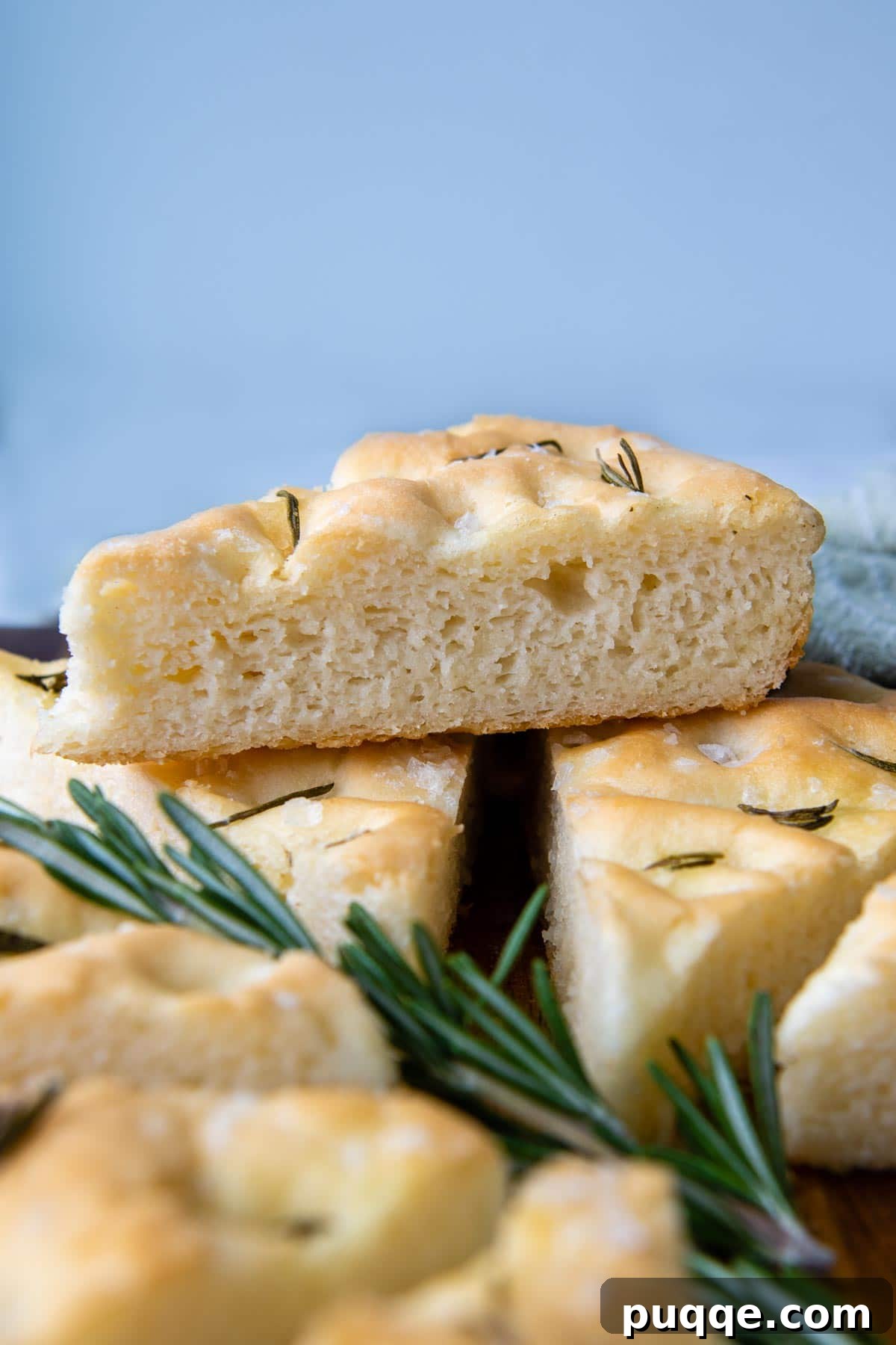 Fluffy Gluten-Free Italian Focaccia Made Easy 2 A golden-brown slice of gluten-free focaccia, resting delicately on a bed of fresh rosemary sprigs, with more slices of the rustic Italian bread stacked in the background.