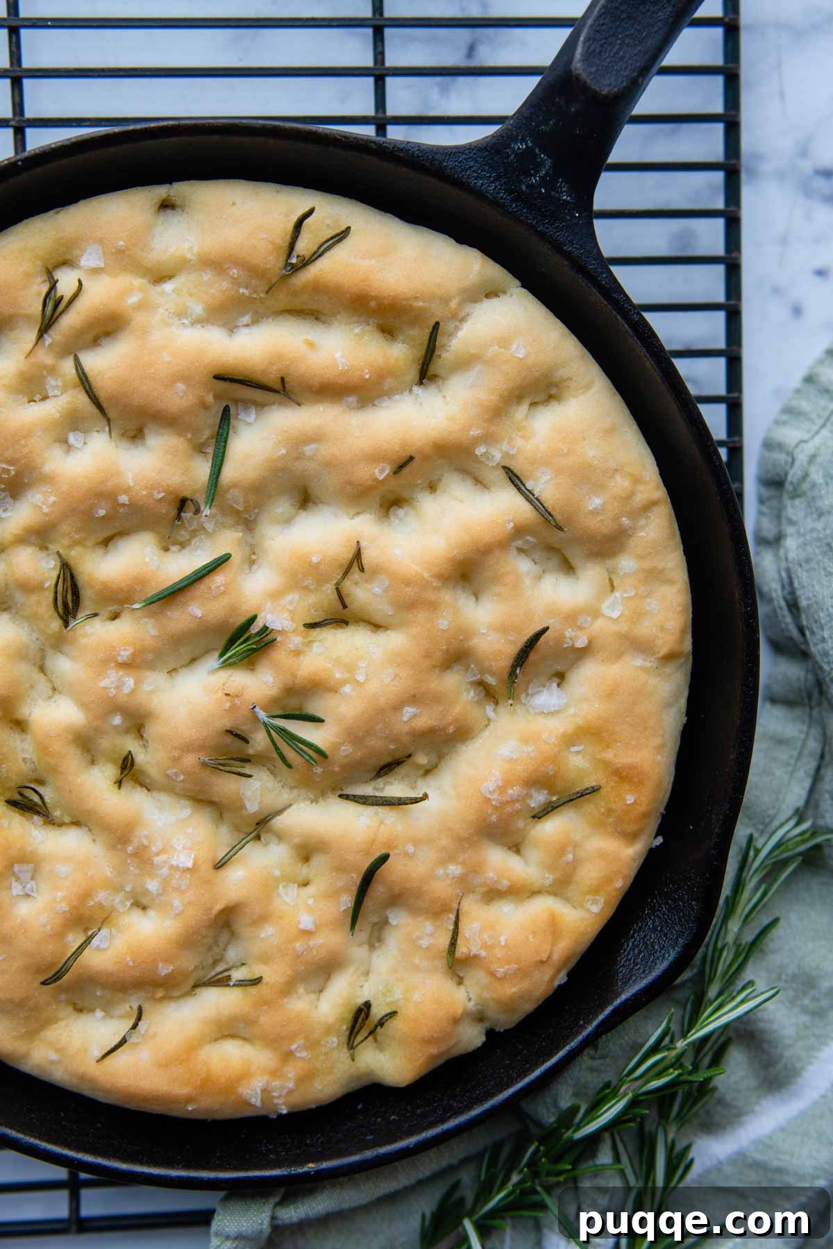 Fluffy Gluten-Free Italian Focaccia Made Easy 7 A freshly baked, unsliced gluten-free focaccia bread resting in a cast iron skillet on a wire cooling rack, showcasing its golden-brown crust and dimpled surface.