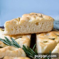 A golden-brown slice of gluten-free focaccia, resting delicately on a bed of fresh rosemary sprigs, with more slices of the rustic Italian bread stacked in the background.