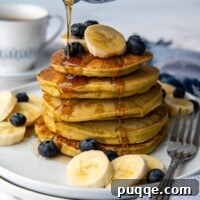 syrup being poured on a stack of pancakes with bananas and blueberries scattered around.