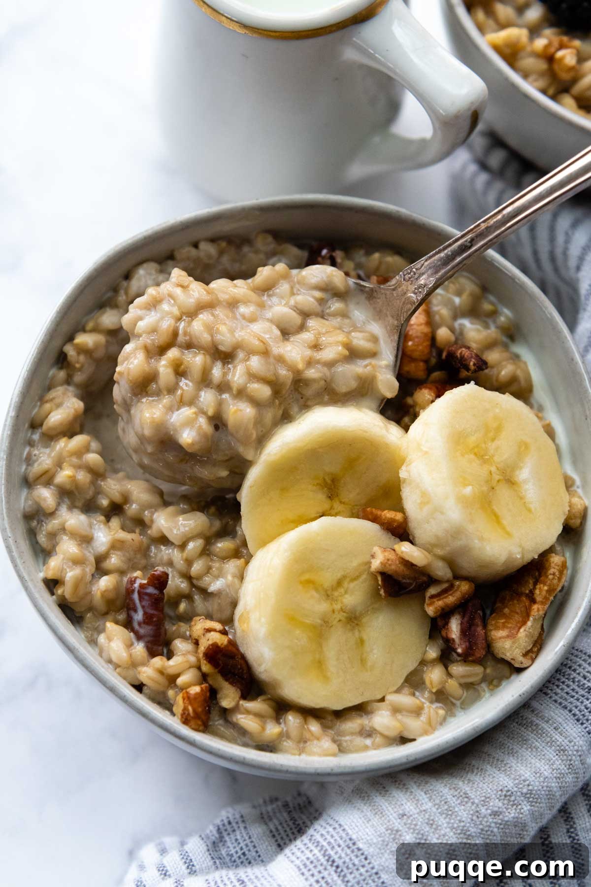 The Perfect Bowl of Creamy, Tender Oat Groats 2 A spoon lifting out creamy, cooked oat groats from a white bowl, garnished with sliced banana and crunchy nuts.