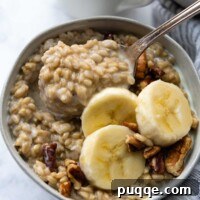 The Perfect Bowl of Creamy, Tender Oat Groats 6 A spoon lifting out creamy, cooked oat groats from a white bowl, garnished with sliced banana and crunchy nuts.