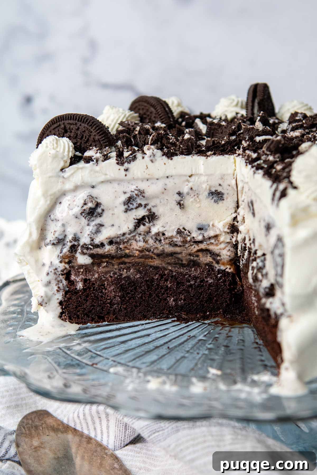 A decadent gluten-free Oreo ice cream cake on a cake stand with a slice removed, revealing layers of chocolate cake, hot fudge, crushed Oreos, and ice cream.