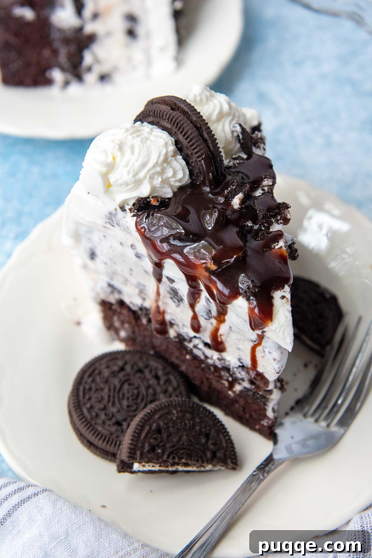 A close-up view of the top of a slice of gluten-free ice cream cake, showing rich hot fudge drizzled over the whipped cream and crushed Oreos.