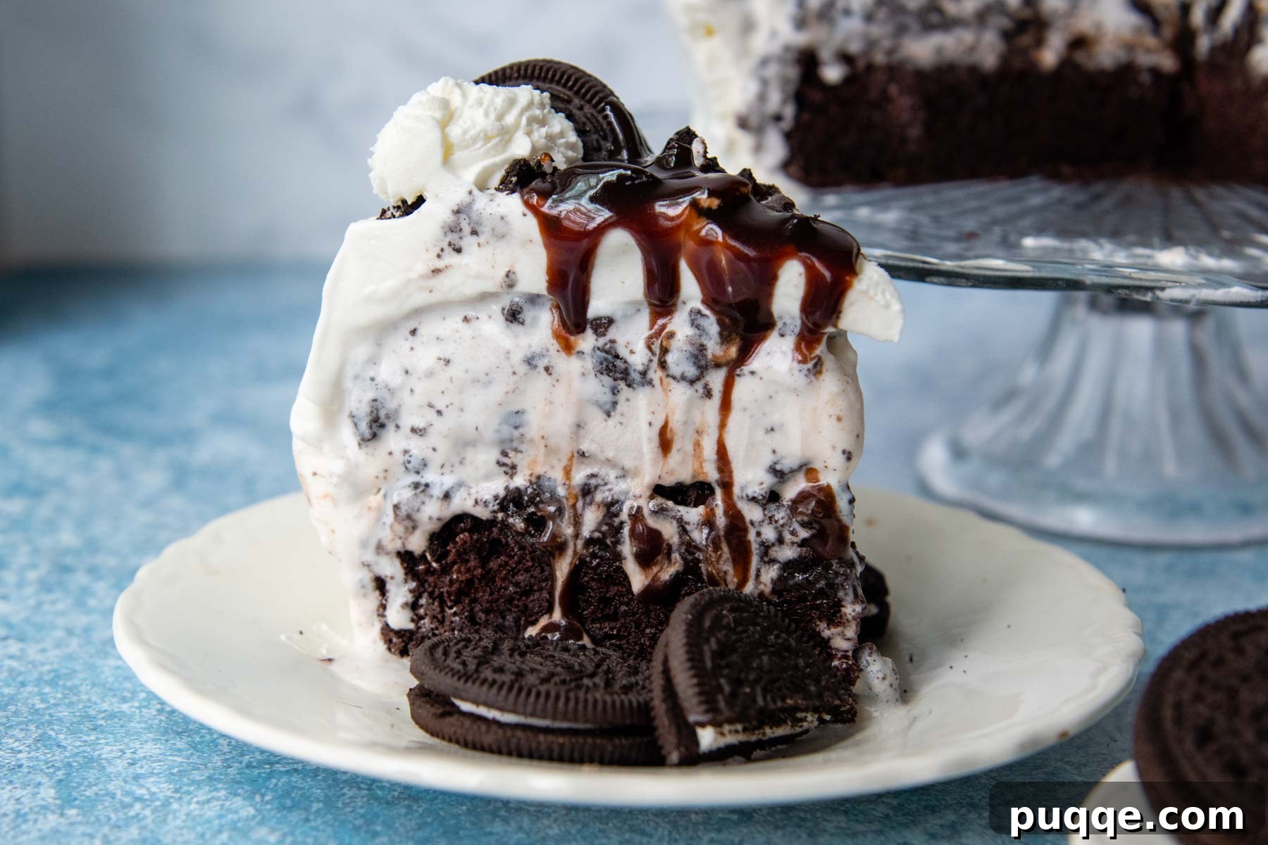 A perfectly portioned slice of ice cream cake, adorned with crushed Oreos and a generous drizzle of homemade hot fudge sauce, presented beautifully on a white plate.