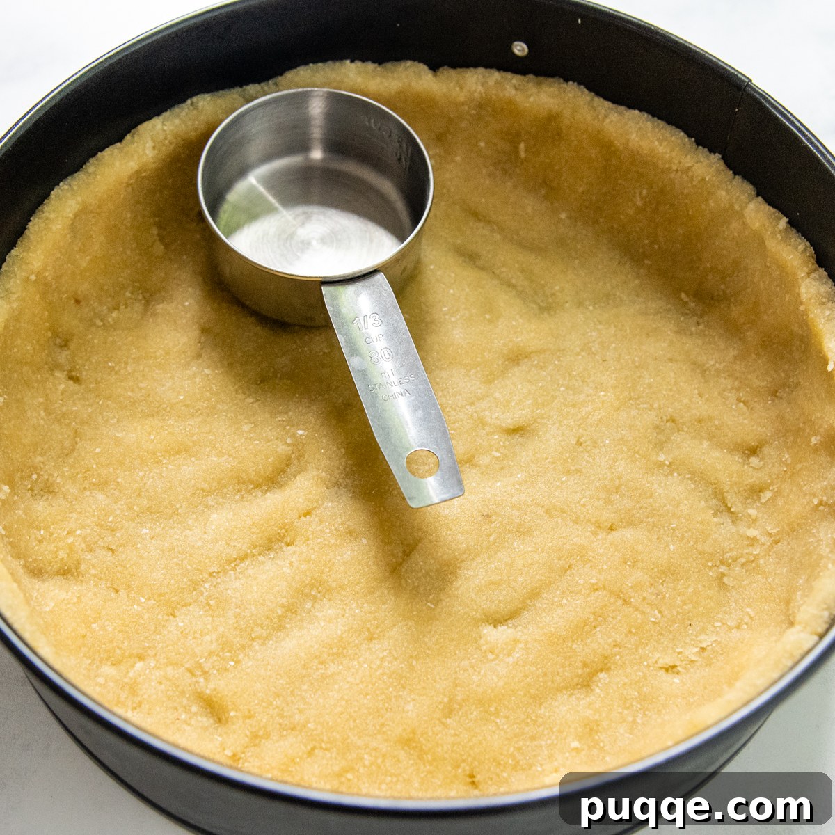 Quick Dreamy Gluten Free No Bake Cheesecake 3 A measuring cup pressing an almond flour crust into a 9-inch springform pan, creating an even, firm base.