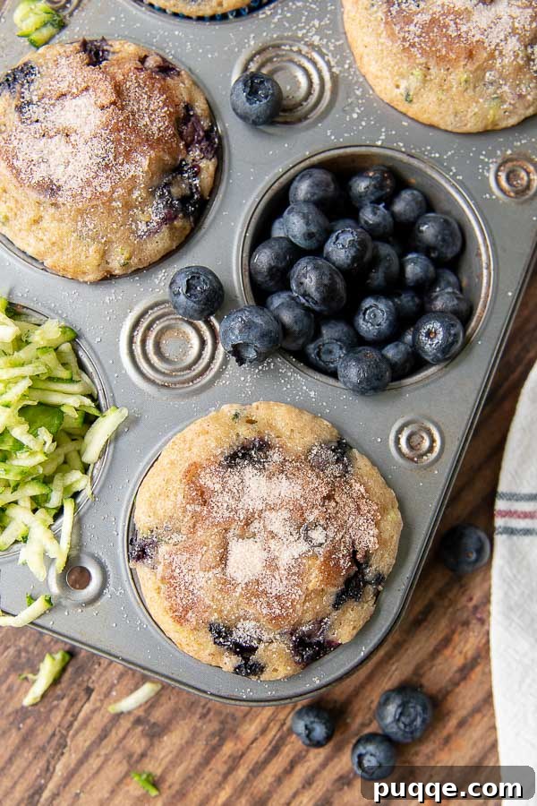 Perfectly Moist Blueberry Zucchini Bread 6 Blueberry zucchini muffins cooling in a tin, with fresh blueberries and shredded zucchini scattered around.