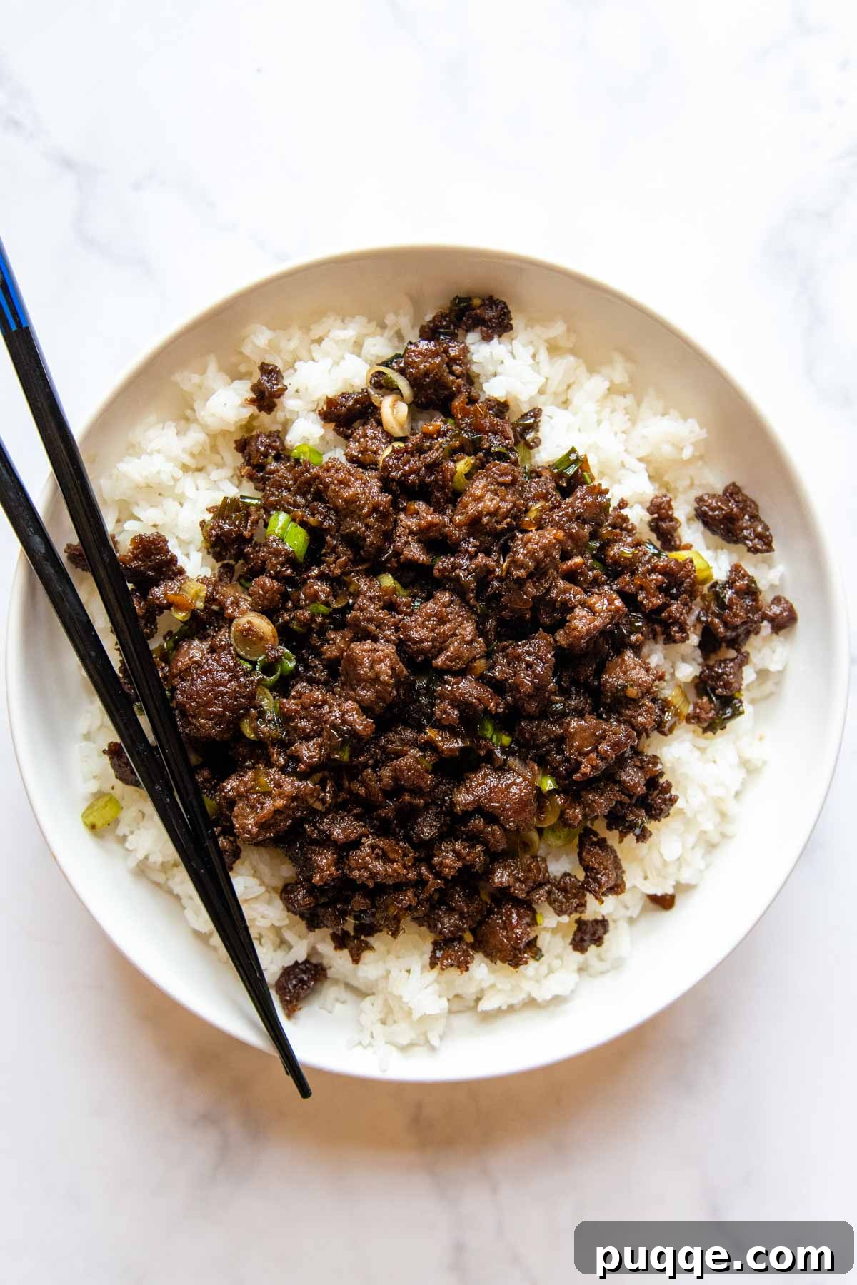 Flavorful Gluten-Free Korean Ground Beef Bulgogi 5 overhead shot of bulgogi served over rice with chopsticks on a white background, garnished with fresh toppings.