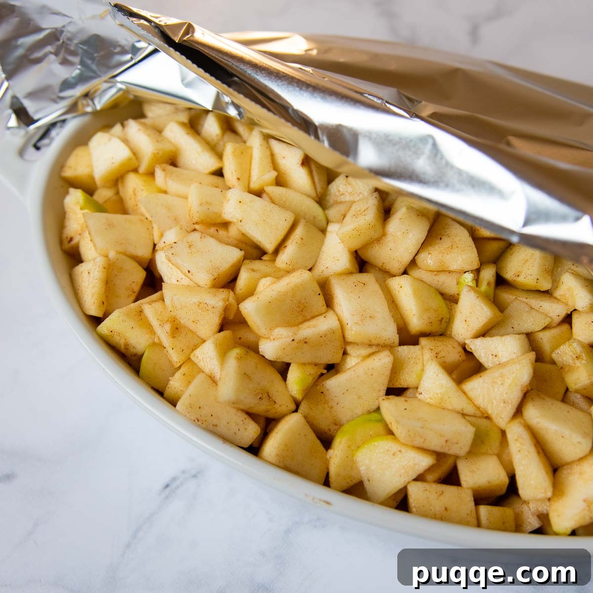 Unbaked apple filling mixture in a baking dish, covered with foil, ready for its initial bake.