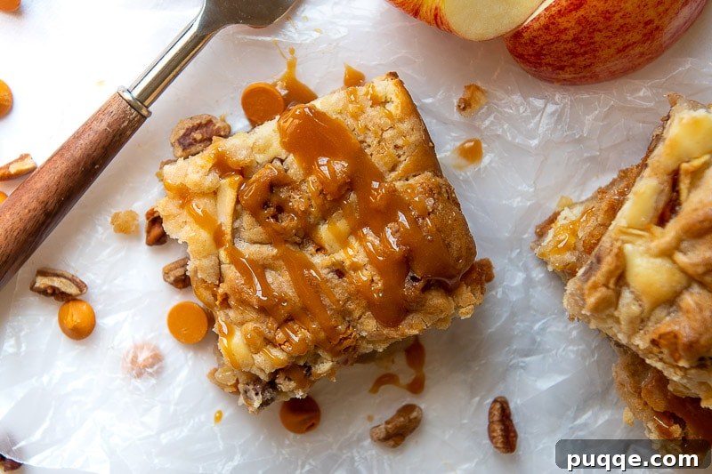 A perfectly sliced caramel apple blondie cheesecake bar resting on wax paper, with a generous drizzle of caramel sauce over its rich layers.