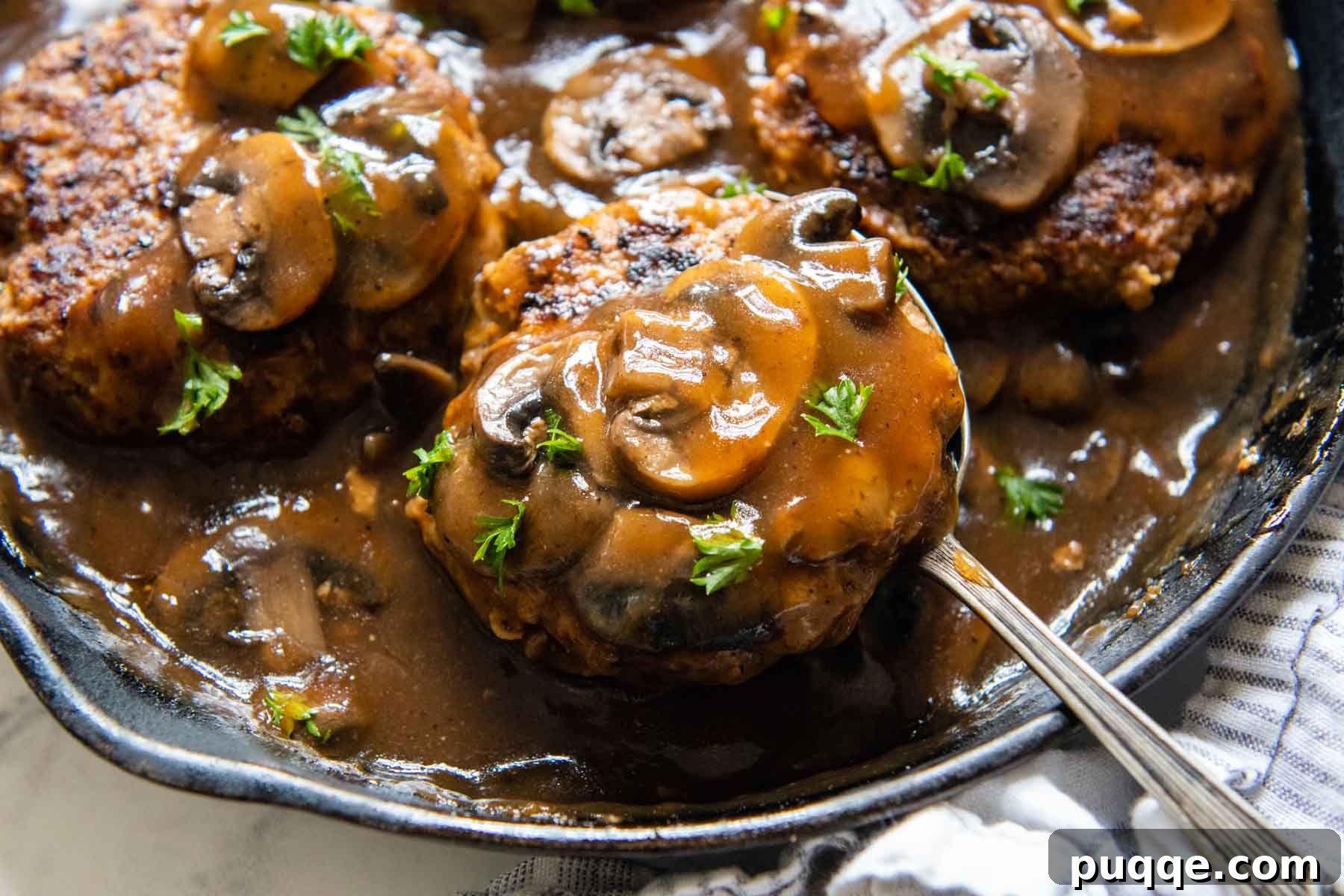 A spoon delicately lifting a perfectly cooked Salisbury steak, adorned with savory mushrooms and gravy, from its skillet.