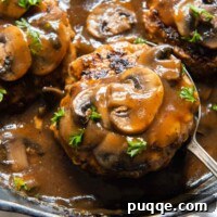 a spoon lifting up a salisbury steak with mushrooms on top from a skillet.