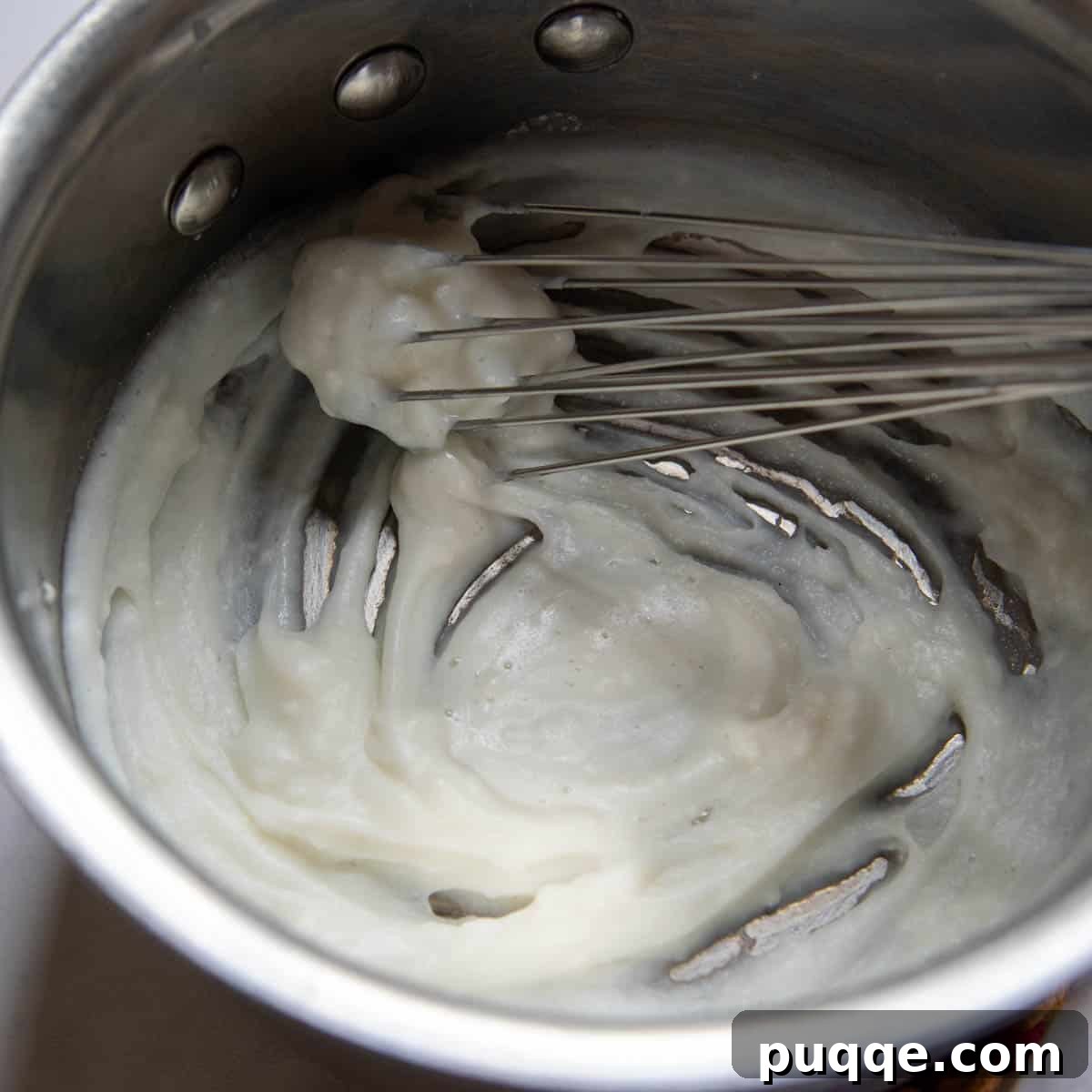 A small saucepan containing the white, thickening roux, with a whisk creating a trail at the bottom.