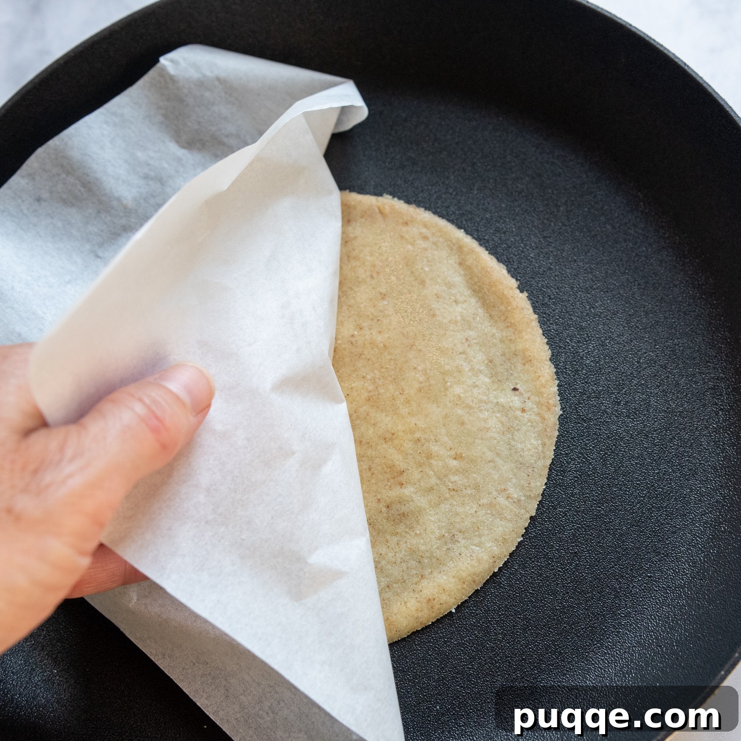 The parchment paper being gently lifted off a tortilla as it cooks in a hot skillet.