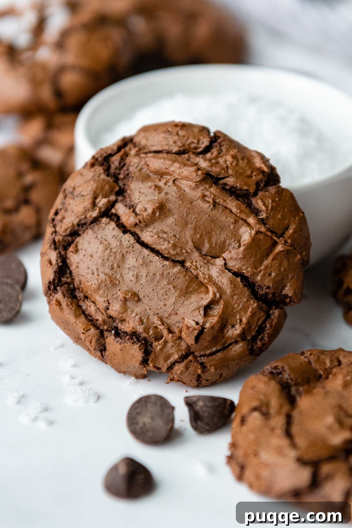 Effortless Gooey Gluten Free Brownie Cookies 7 A single fudgy gluten-free brownie cookie, perfectly cracked on top, rests invitingly against a small white bowl of flaky sea salt, highlighting its delicious texture.