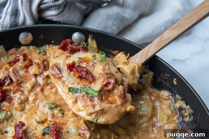 Pratik Kremalı Kurutulmuş Domatesli Enginarlı Tavuk 7 Closer angle of a spoon lifting a piece of creamy sun-dried tomato chicken with sauce and herbs from the skillet.