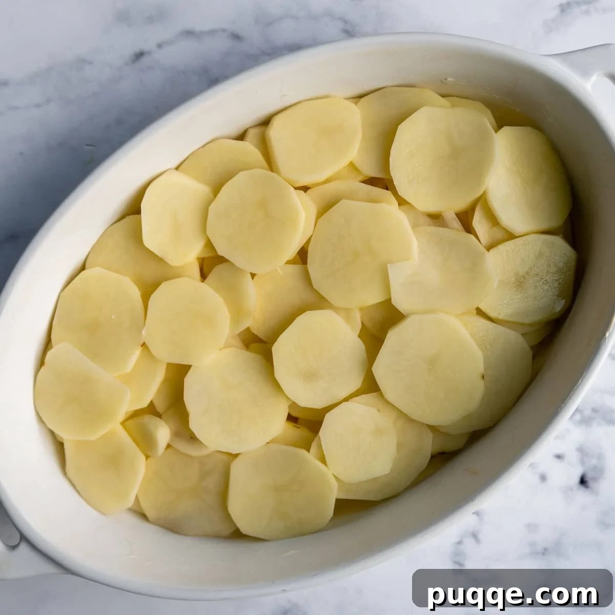A layer of thinly sliced, unbaked russet potatoes neatly arranged in a greased baking dish, ready for the creamy sauce.