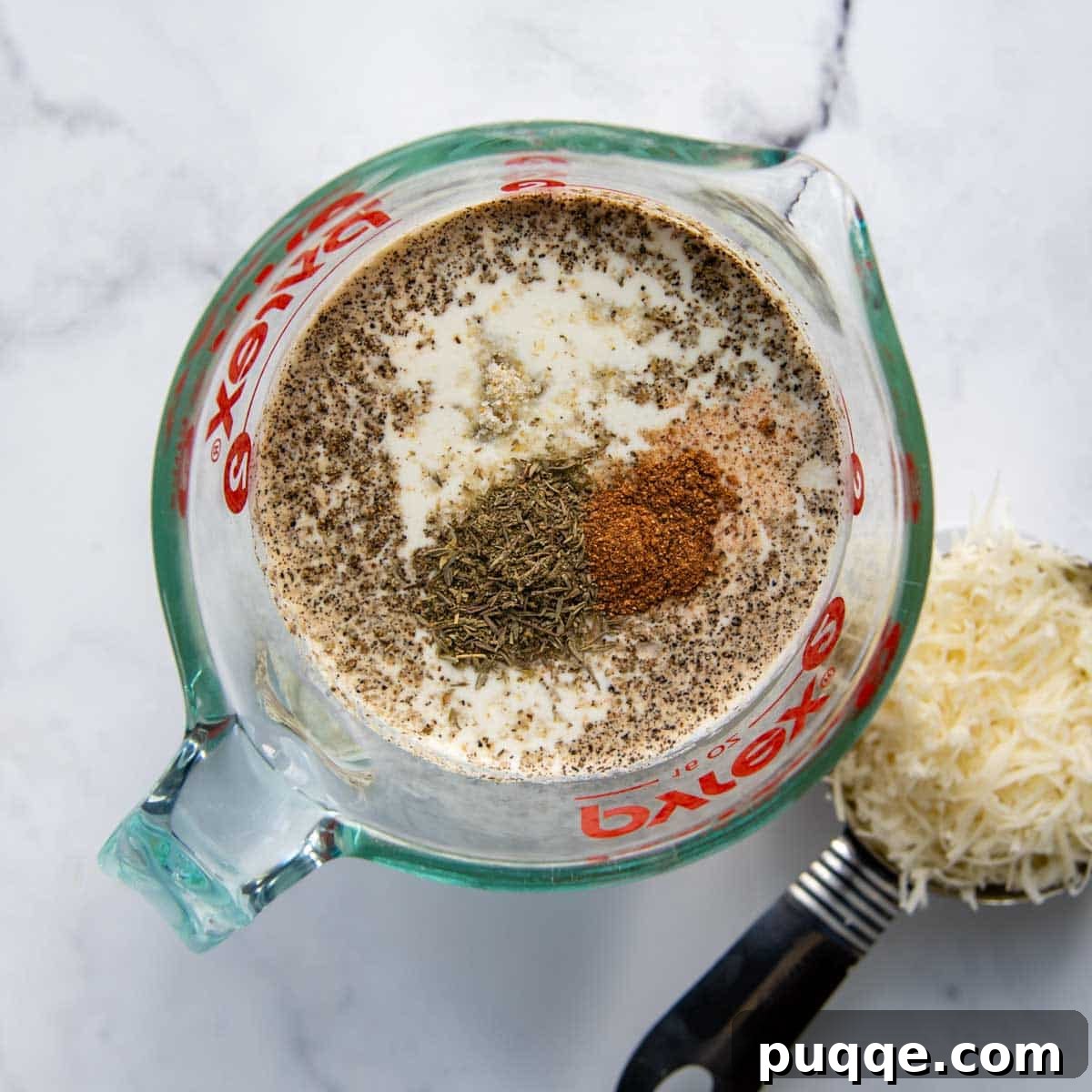 A bowl of creamy, seasoned sauce with a whisk, next to piles of freshly shredded Swiss and Parmesan cheeses, ready for layering.