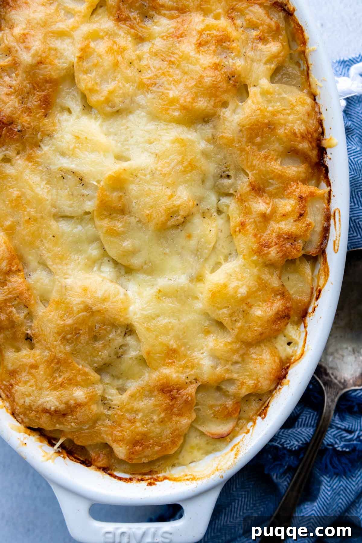An overhead shot of a white casserole dish filled with unbaked au gratin potatoes, with a blue kitchen towel draped nearby.
