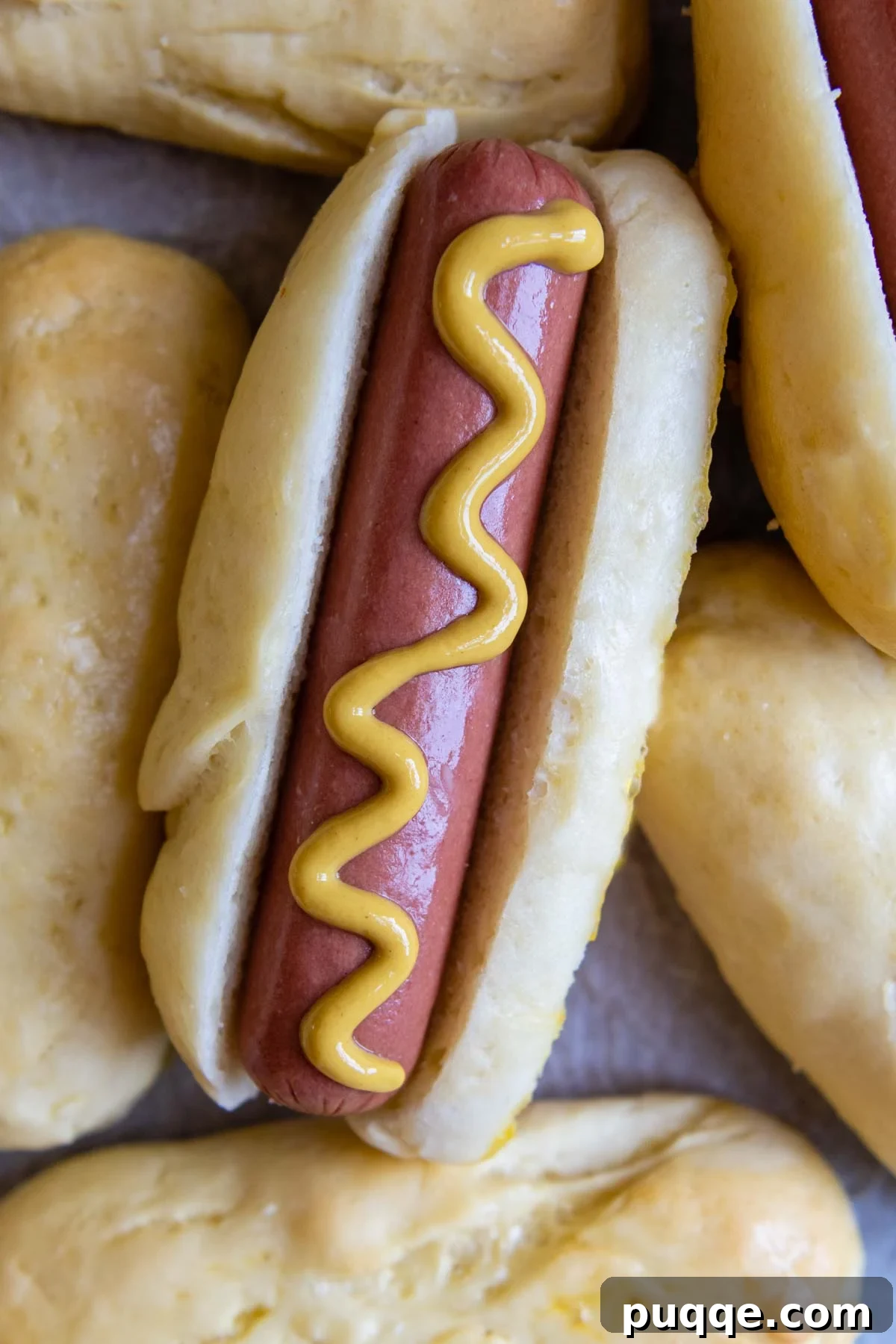 A perfectly grilled hot dog nestled in a homemade gluten-free bun, drizzled with mustard, next to a stack of fresh buns.