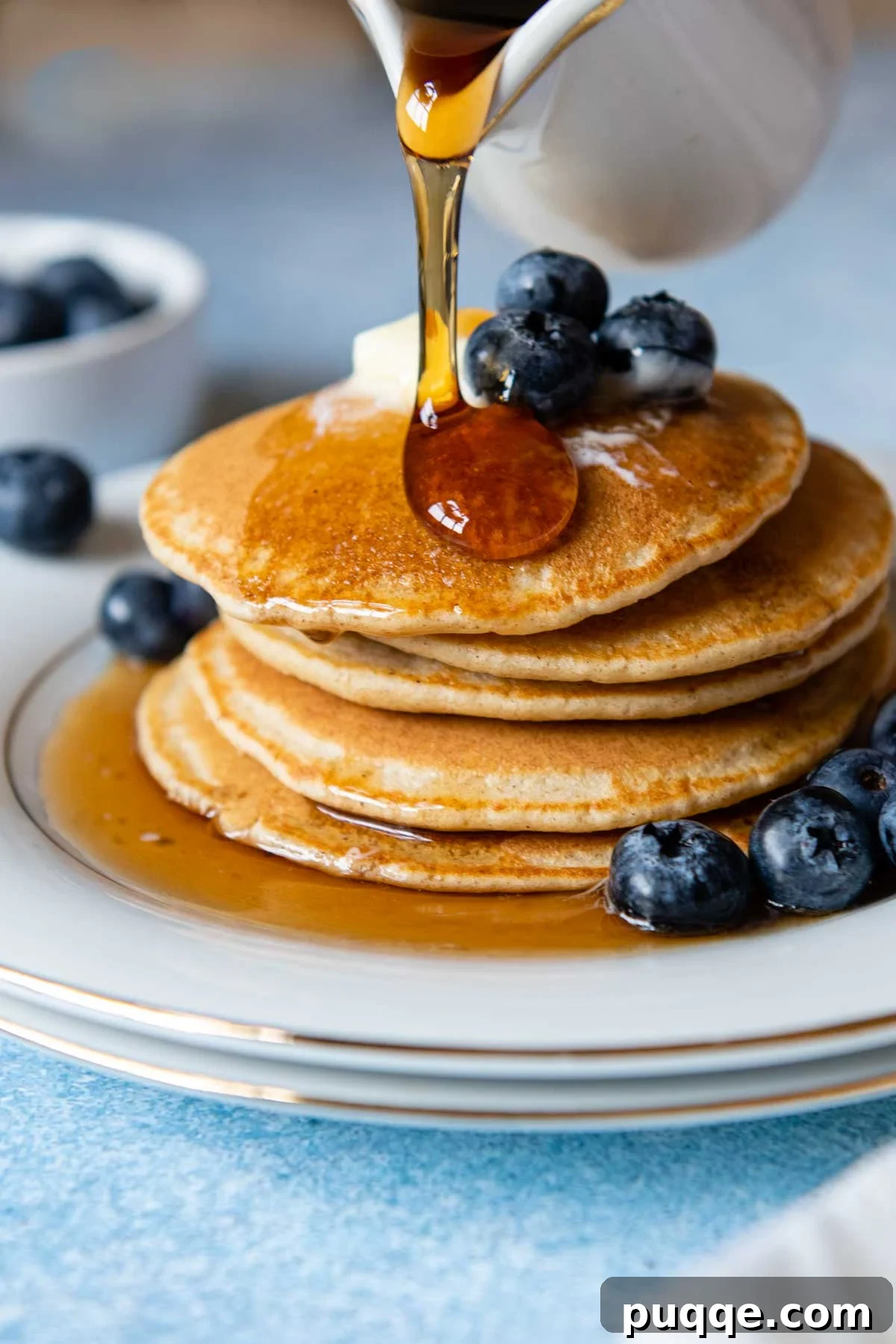Golden syrup being poured generously over a beautifully stacked tower of fluffy oat flour pancakes, with fresh berries scattered around.