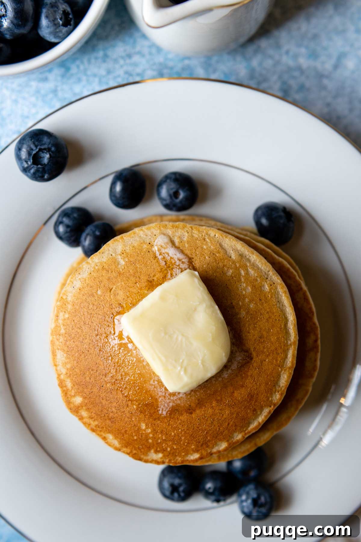 A beautiful stack of golden-brown oat flour pancakes on a white plate, adorned with a scattering of fresh blueberries.
