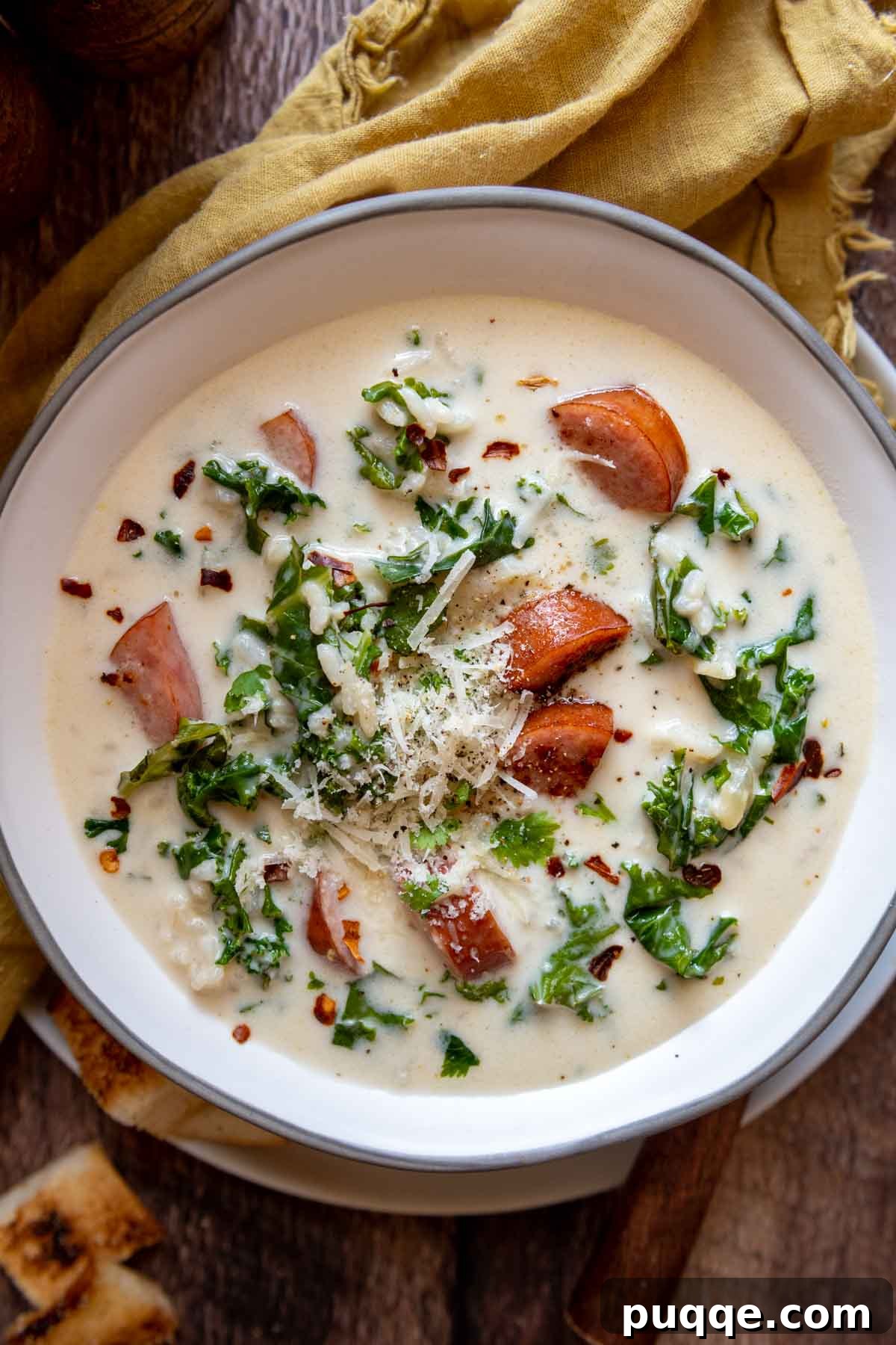 Overhead shot of creamy kale and sausage soup in a white bowl, with a yellow napkin beside it.