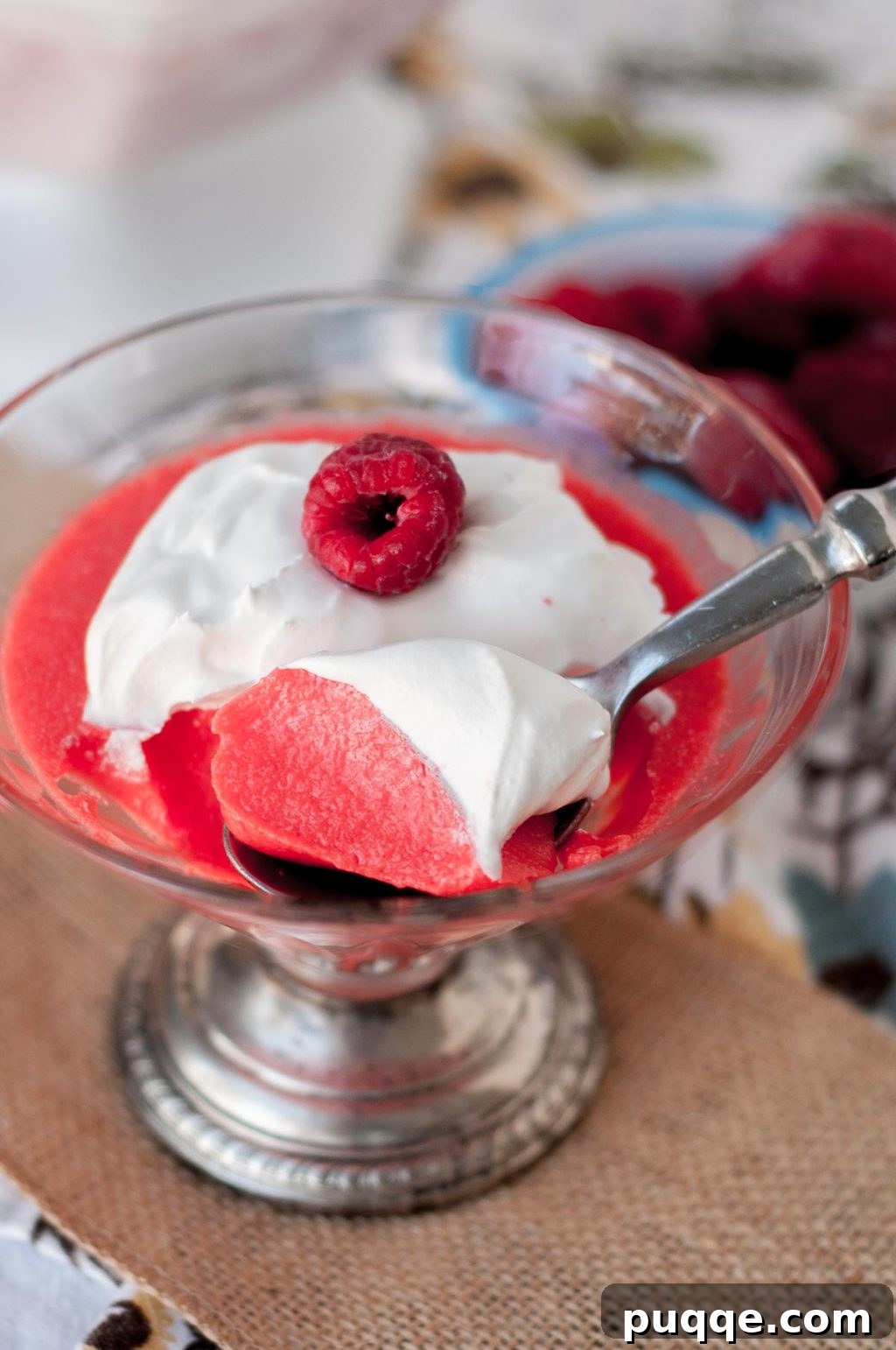 A spoon scooping out a bite of creamy raspberry Jello, with a dollop of whipped cream on top, in a glass bowl.