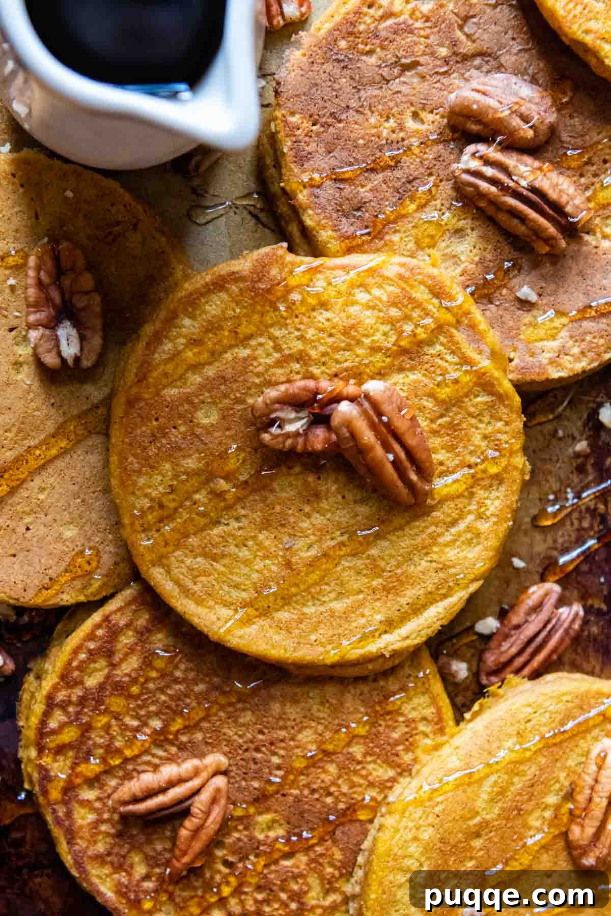 Overhead shot of fluffy pumpkin oatmeal pancakes on a pan, drizzled with syrup and topped with nuts, highlighting their golden-brown perfection.