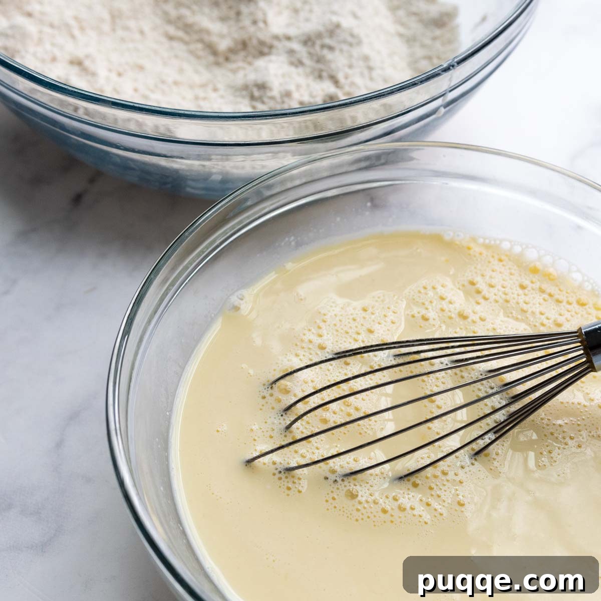 Two bowls side-by-side on a countertop; one contains whisked wet ingredients (milk, eggs, butter, vanilla), and the other holds the dry ingredients (flour, sugar, leavening agents).