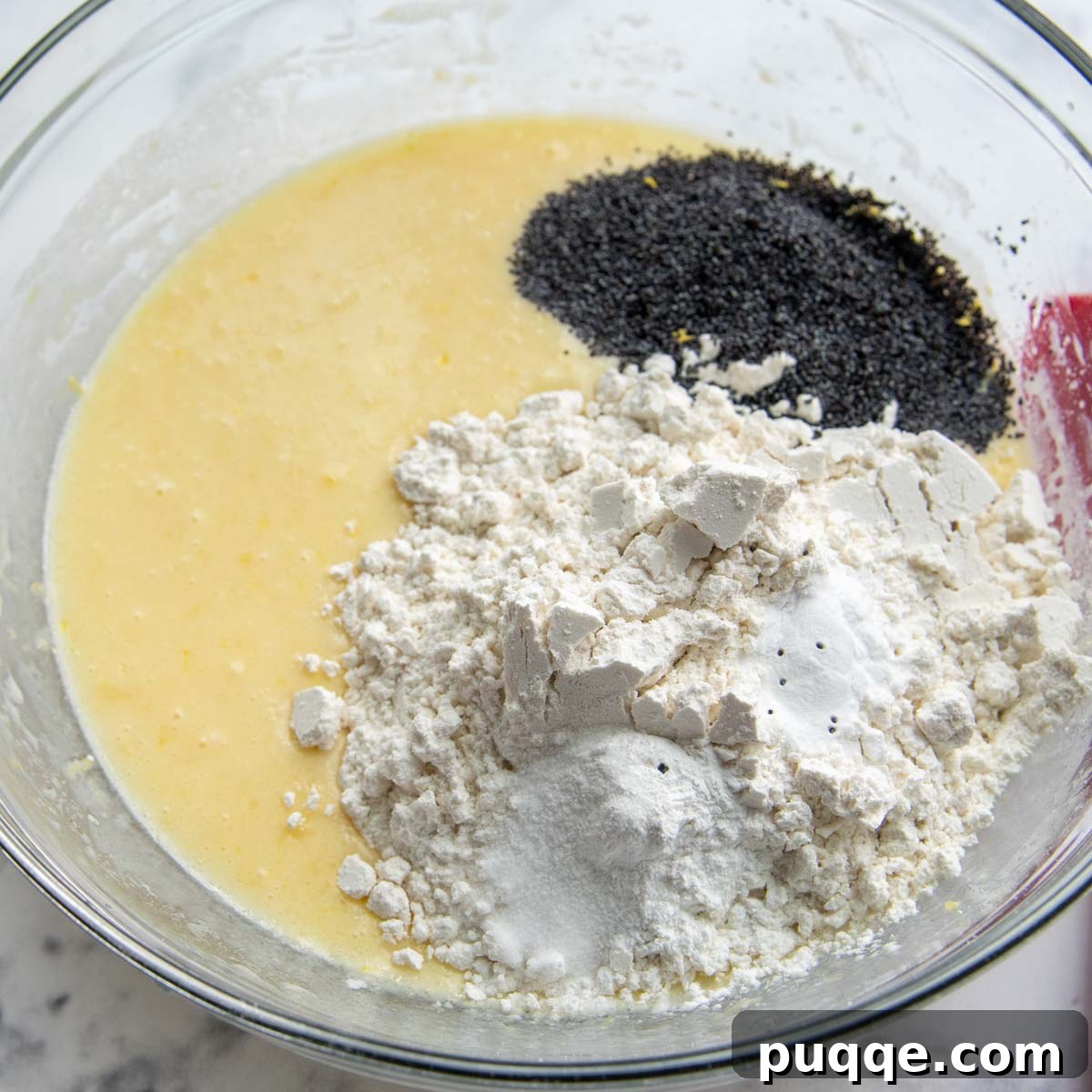 Muffin batter in a glass bowl, with gluten-free flour and poppy seeds ready to be gently mixed in.