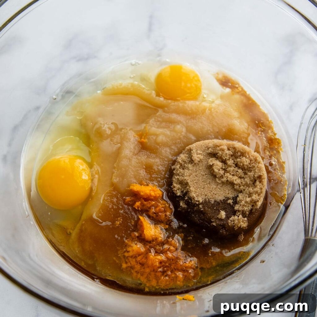 Wet ingredients including eggs, applesauce, oil, and sugar being whisked in a large glass bowl.