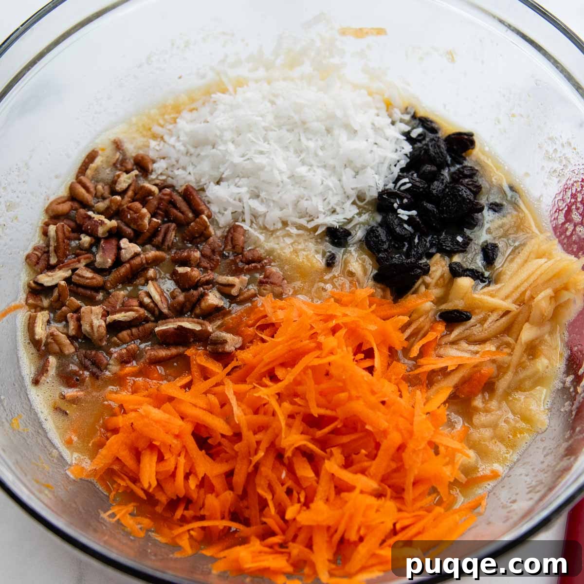 Grated carrots, apples, raisins, coconut, and pecans added to the wet mixture in a bowl.
